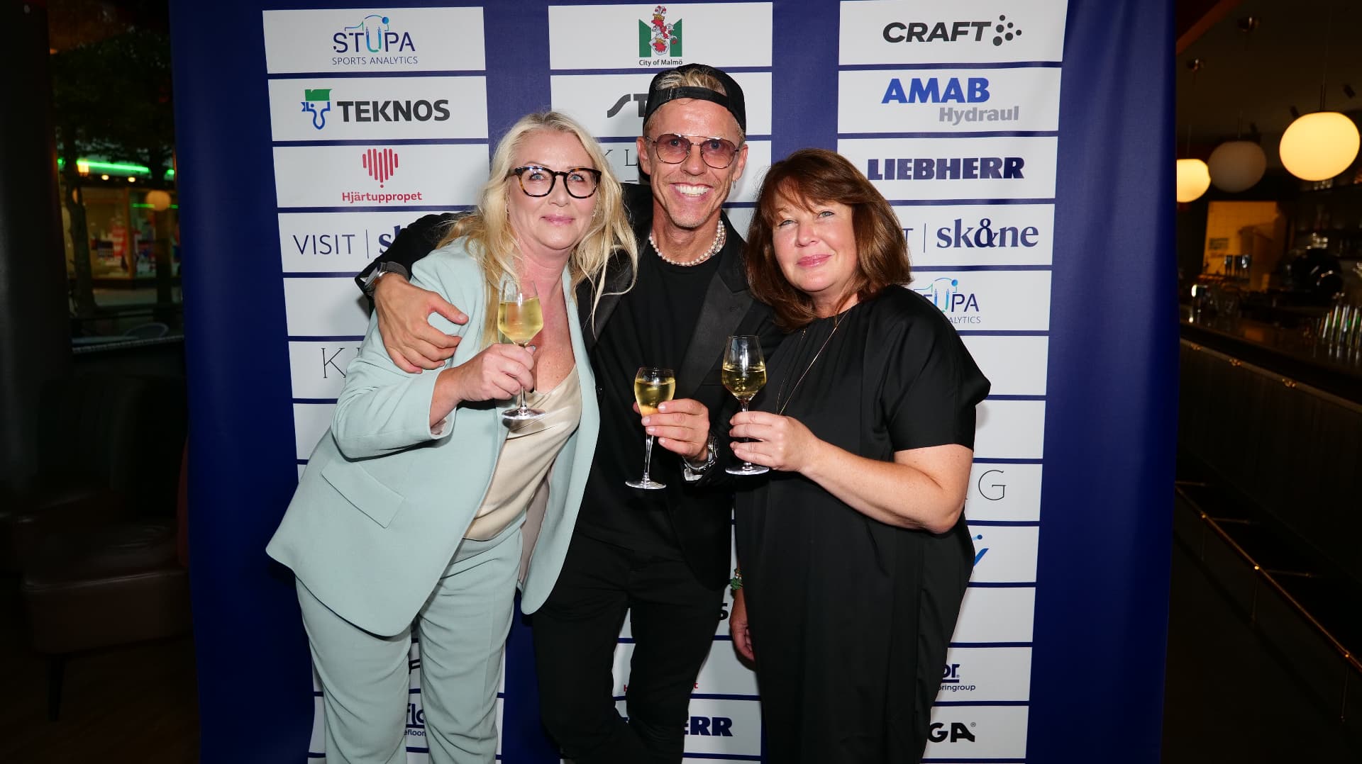 Three guests raise a toast in front of the sponsor wall at the World Champions' Dinner in Malmö